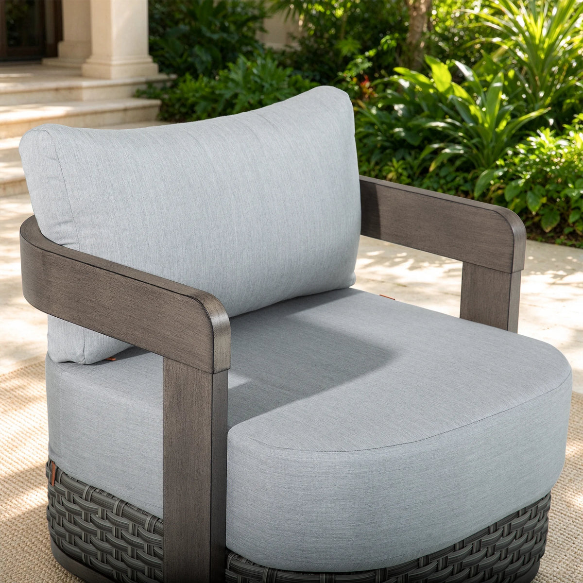 The Manarola modern curved swivel club chair styled on a sunny outdoor patio rug with lush garden greenery in the background.