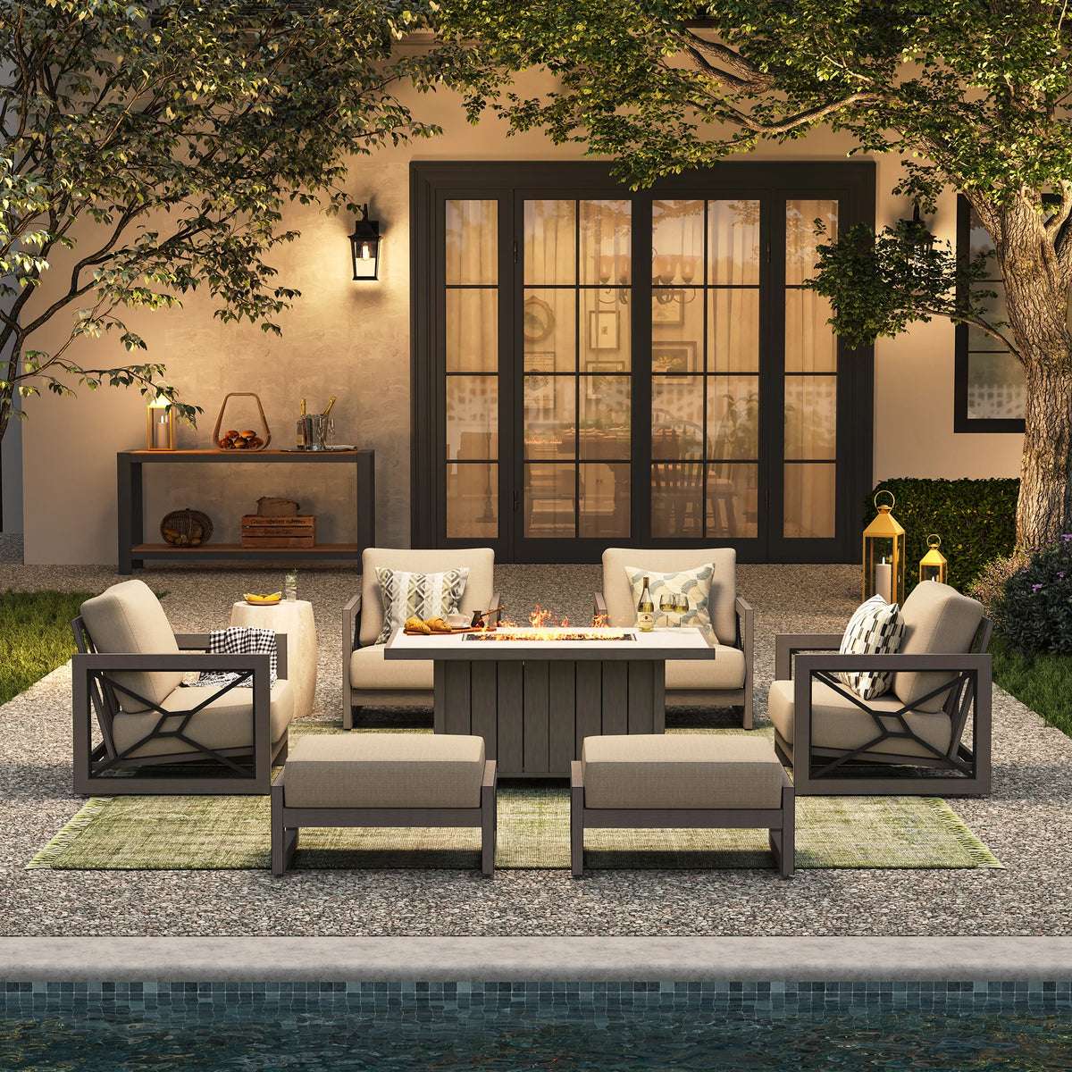 A high-end evening poolside setup offering backyard staycation ideas, with the Marindo 7-piece lounge suite arranged over a green rug with a glowing fire pit and ottomans styled as extra front benches.