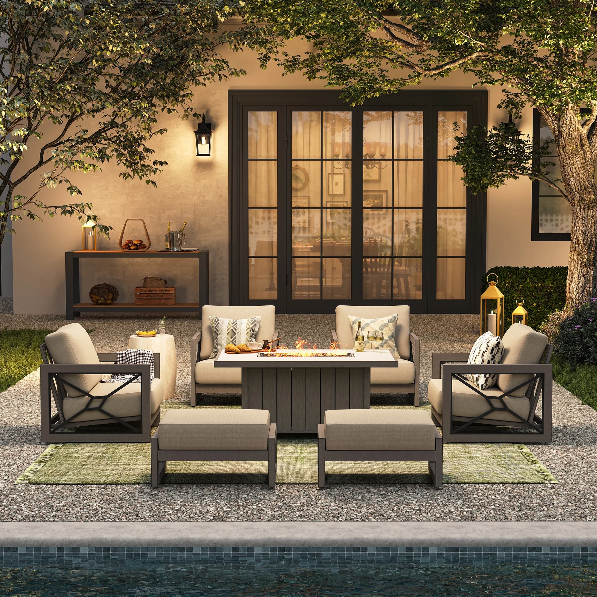 A high-end evening poolside setup offering backyard staycation ideas, with the Marindo 7-piece lounge suite arranged over a green rug with a glowing fire pit and ottomans styled as extra front benches.