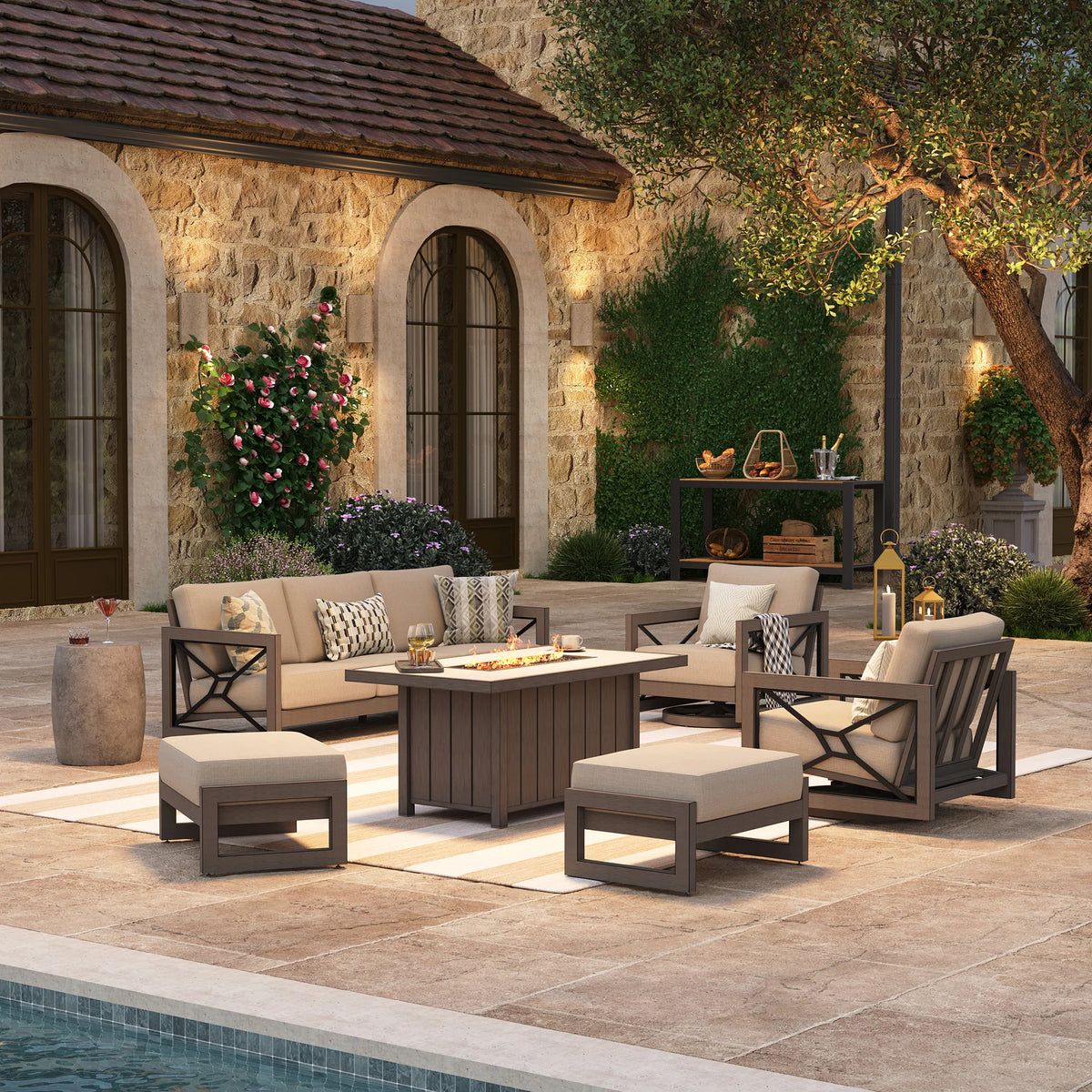 Upscale outdoor entertaining space at night featuring the Marindo 6-piece aluminum seating set, with amber flames glowing from the stainless steel fire pit table surrounded by sofas, swivel chairs, and ottomans.