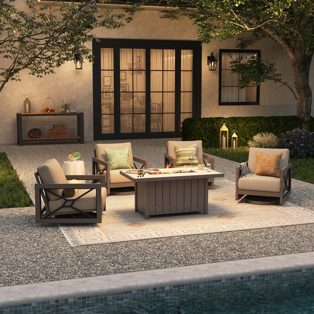 An elegant poolside entertaining space at night, showing the Marindo 5-piece fire chat retreat with glowing amber flames from the propane table throwing warm light onto four swivel lounge chairs.