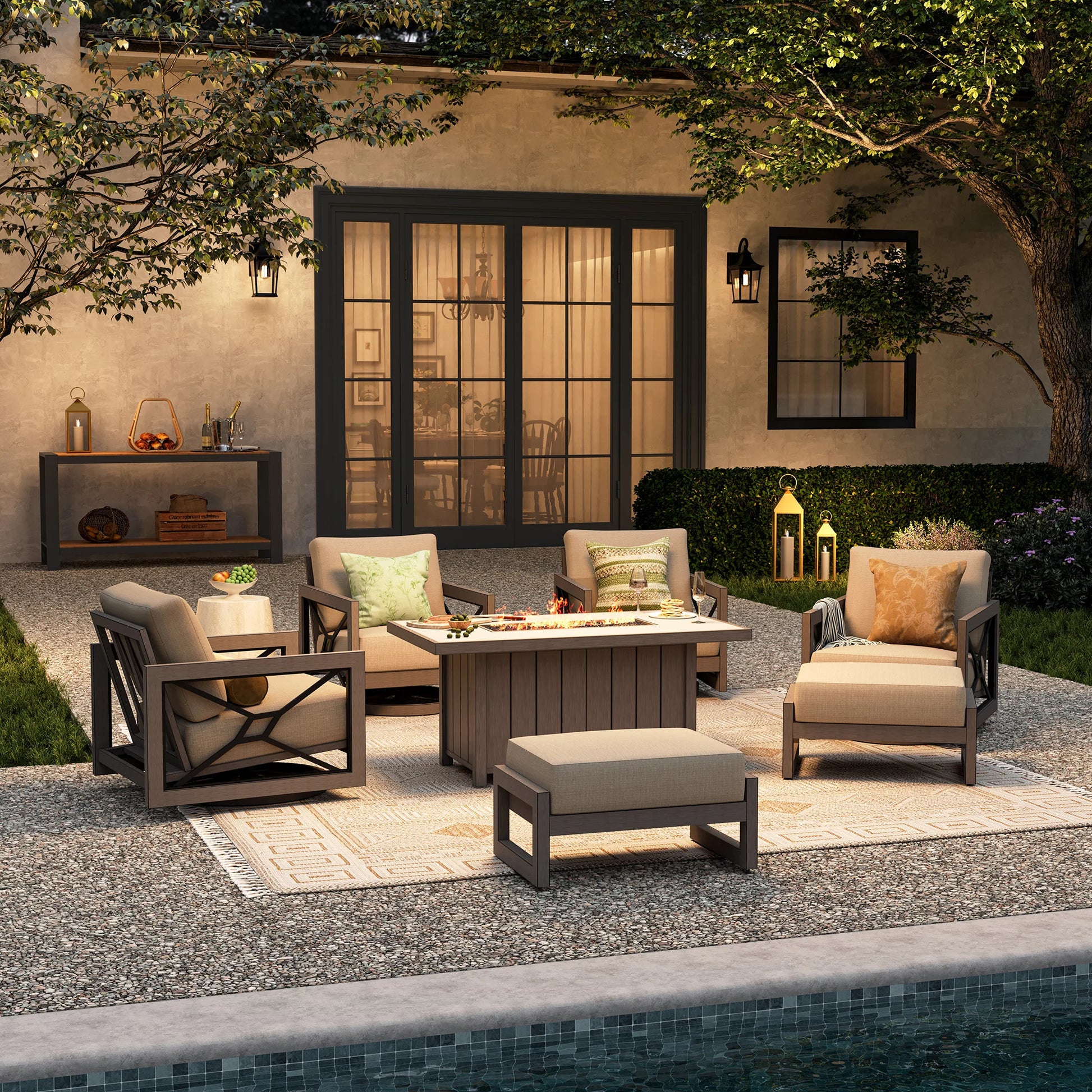 An elegant evening pool deck providing backyard gathering inspiration, displaying the Marindo 7-piece conversational swivel lounge with glowing flames from the fire table and ottomans placed as flexible front benches.