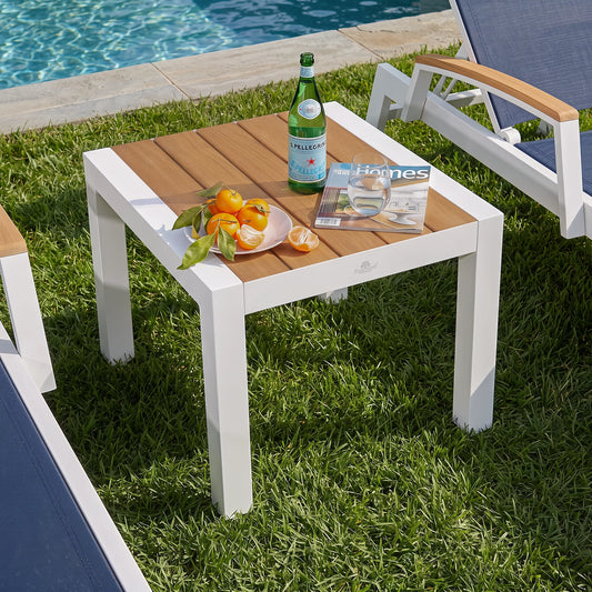 Square outdoor patio side table with white aluminum frame and slatted teak wood tabletop, poolside end table beside lounge chairs on green lawn