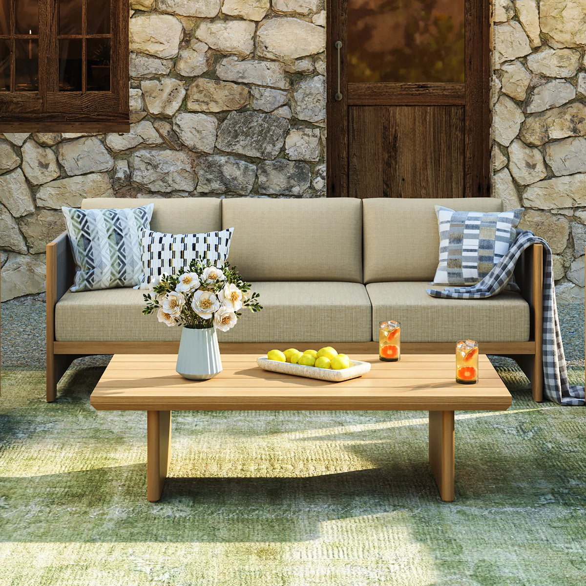 Horizontal view of a slatted faux-wood outdoor coffee table holding yellow lemons and iced drinks in front of a beige sofa and rustic stone cottage wall.
