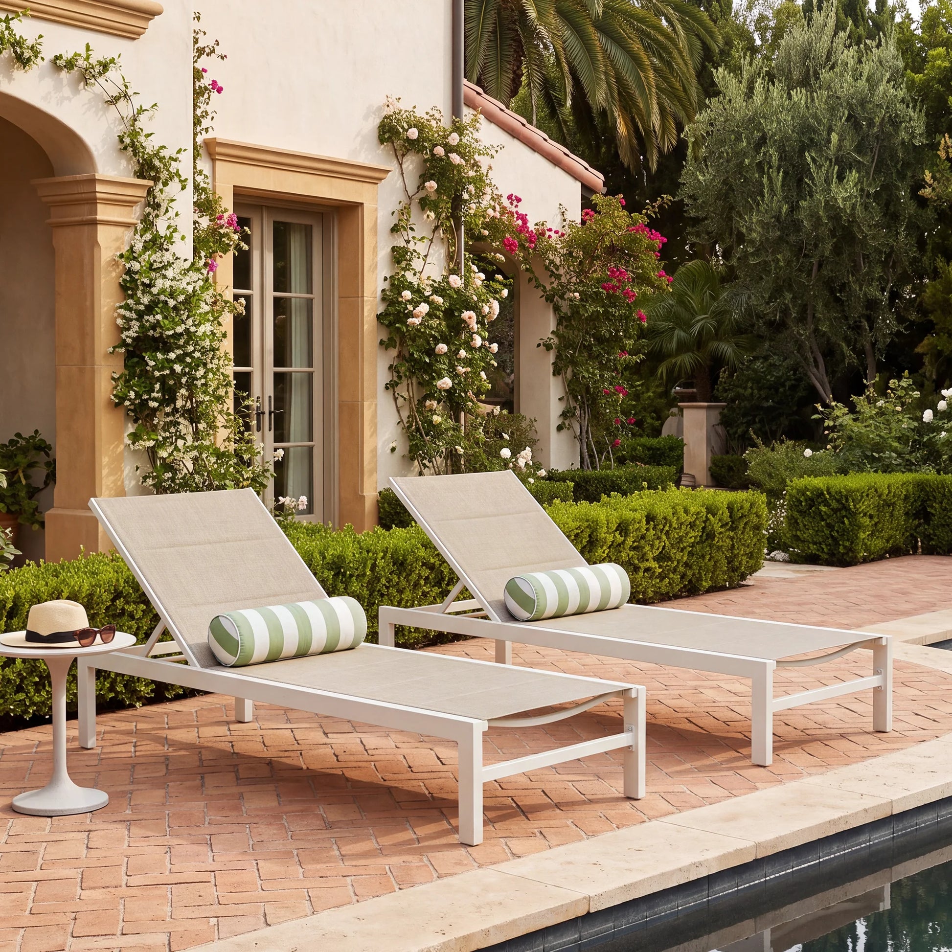 Pair of modern white powder-coated aluminum chaise lounge chairs with beige breathable padded Textilene sling fabric and green striped bolster pillows on a brick patio by the pool.