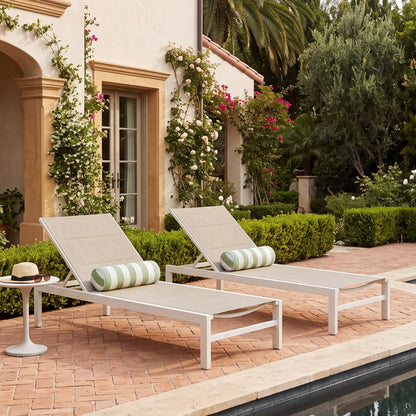 Pair of modern white powder-coated aluminum chaise lounge chairs with beige breathable padded Textilene sling fabric and green striped bolster pillows on a brick patio by the pool.