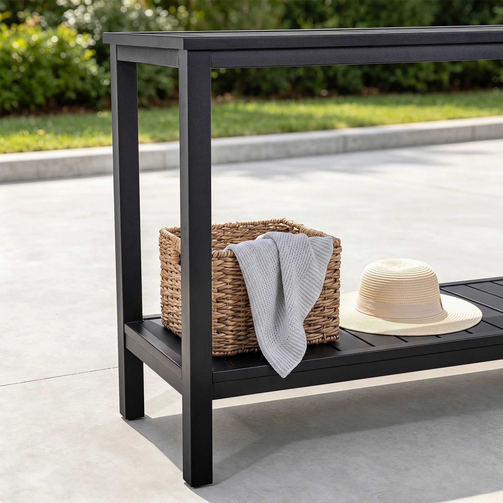 Close-up of Saporta black metal console table bottom shelf featuring a woven storage basket with a gray blanket and a beige straw hat on a concrete patio.