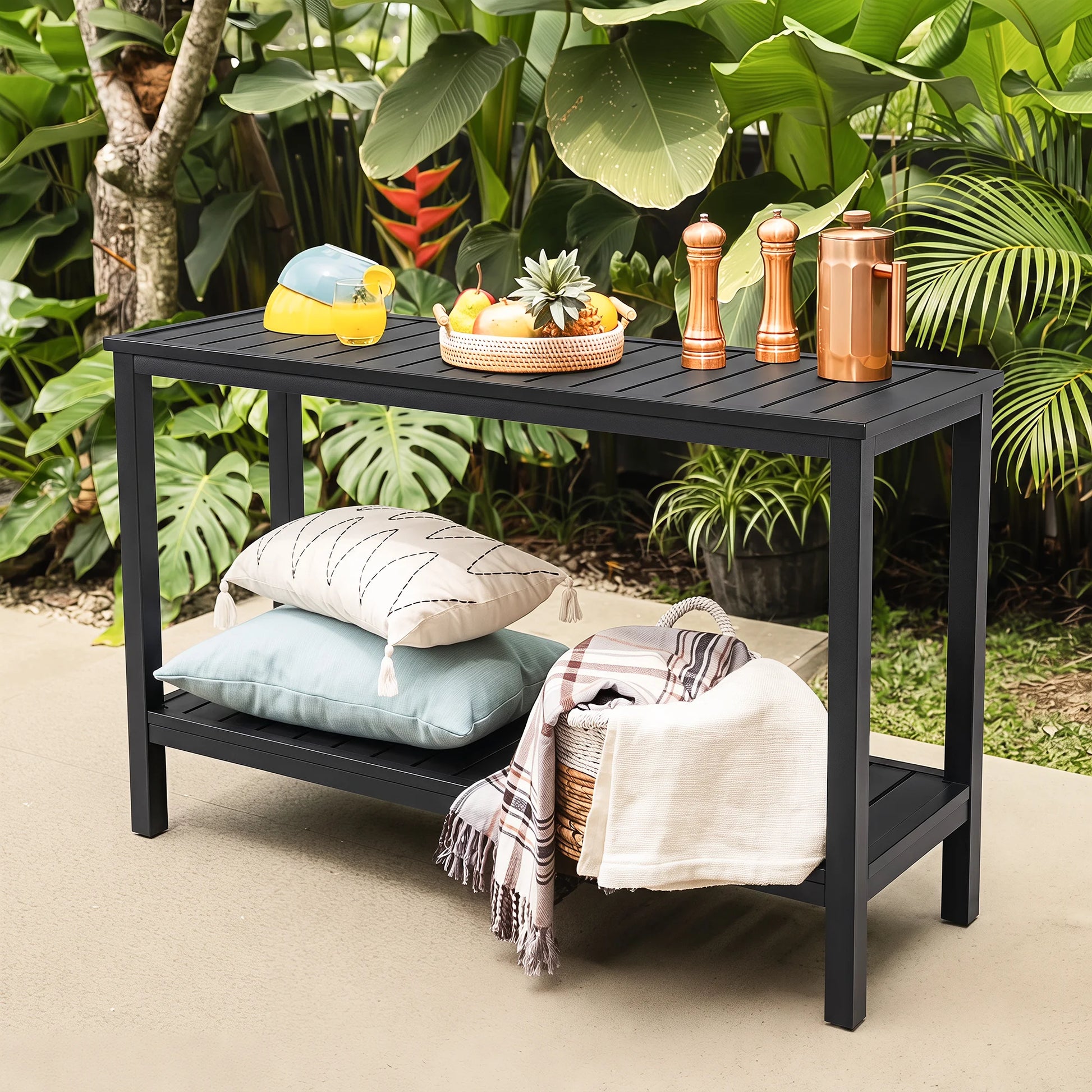 Angled lifestyle shot of Saporta garden serving table styled with a fruit tray, drinks, pillows, and blankets surrounded by lush tropical plants.