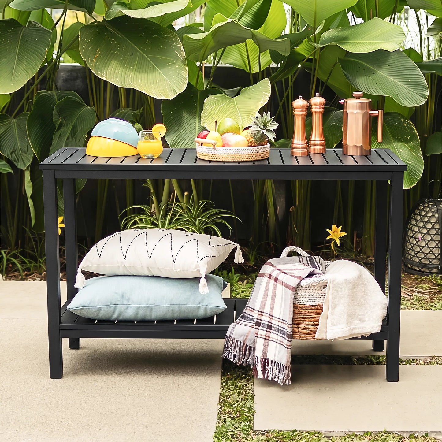 Front view of Saporta outdoor kitchen prep station table with copper pitchers, fresh fruits, and outdoor pillows against a green tropical garden backdrop.