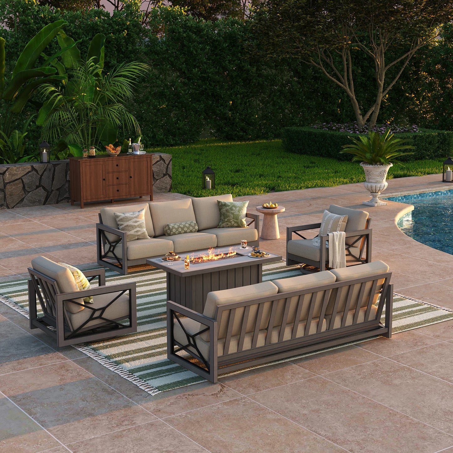 An upscale open-air salon by a pool at sunset, displaying the Marindo 8-guest aluminum patio set with a glowing fire pit centerpiece throwing warm light onto the surrounding sofas and swivel accent chairs.